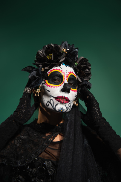Woman with sugar scull halloween makeup touching face isolated on green  - Photo, Image