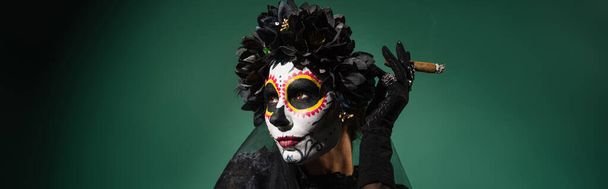 Γυναίκα με santa muerte μακιγιάζ και στεφάνι κρατώντας πούρο σε πράσινο φόντο, πανό  - Φωτογραφία, εικόνα