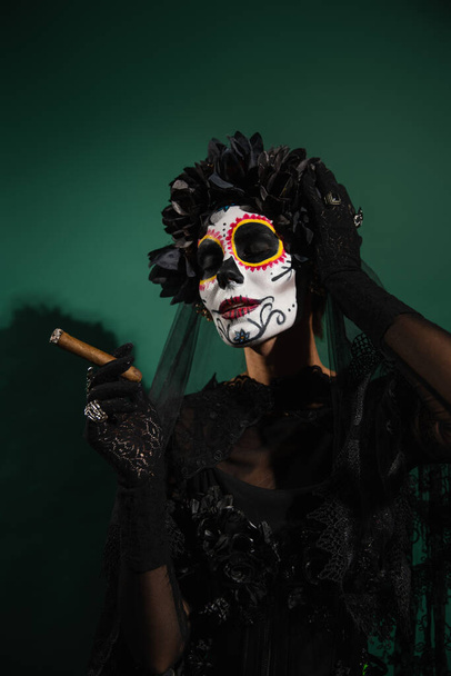 Frau in mexikanischem Totenschminke und Kostüm mit Zigarre auf grünem Hintergrund  - Foto, Bild