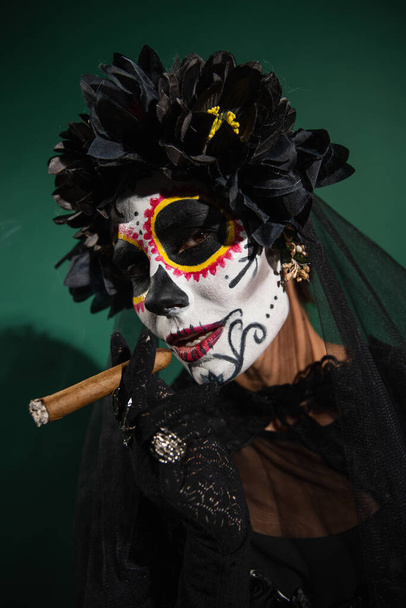 Portrait of woman in santa muerte halloween costume holding cigar on green background  - Zdjęcie, obraz
