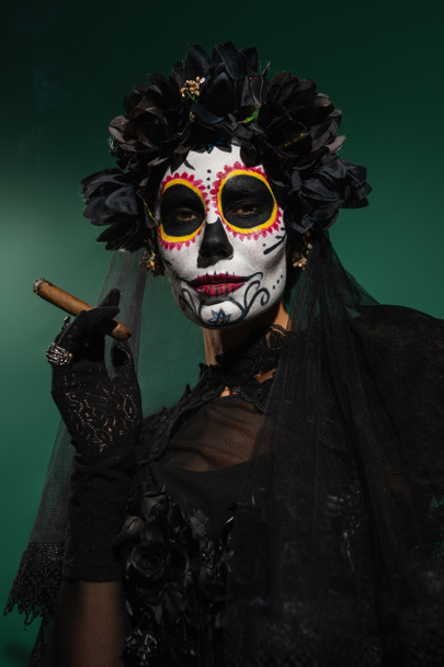 Γυναίκα σε μεξικανική ημέρα του θανάτου μακιγιάζ και μαύρο κοστούμι κρατώντας πούρο στο πράσινο φόντο  - Φωτογραφία, εικόνα