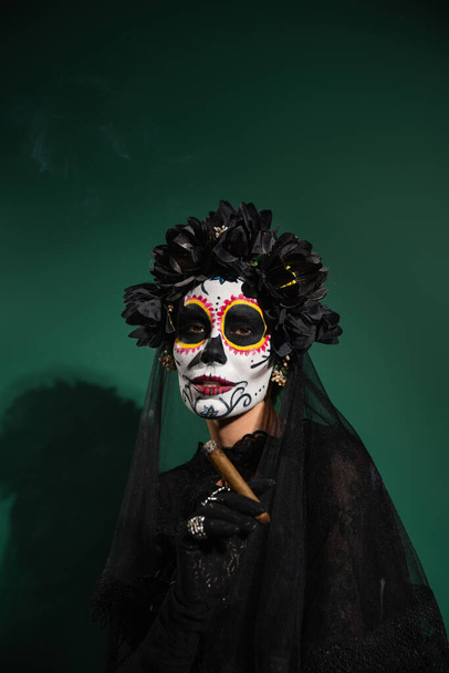 Santa Muerte Cadılar Bayramı kostümü ve yeşil arka planda puro tutan çelenkli kadın.  - Fotoğraf, Görsel