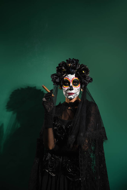 vrouw in santa muerte halloween kostuum houden sigaar op groene achtergrond  - Foto, afbeelding