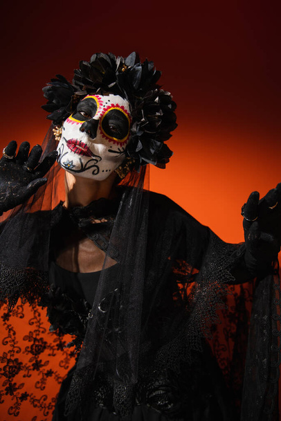 Woman with spooky sugar skull makeup and black costume standing on red  - Fotografie, Obrázek