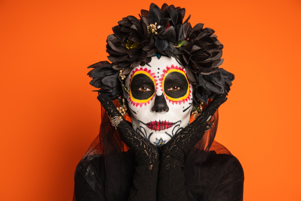 portrait of woman in black wreath and sugar skull halloween makeup posing with hands near face isolated on orange - Photo, Image