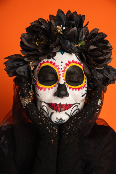retrato de mulher sorridente com olhos fechados e maquiagem assustadora halloween segurando as mãos perto do rosto isolado na laranja - Foto, Imagem
