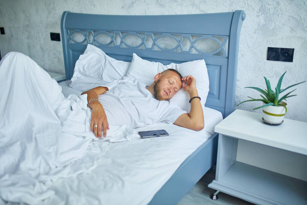 Homme barbu dormant seul sur un grand et confortable linge de lit blanc avec alarme smartphone à la maison, mode de vie, oreiller confortable, lit - Photo, image