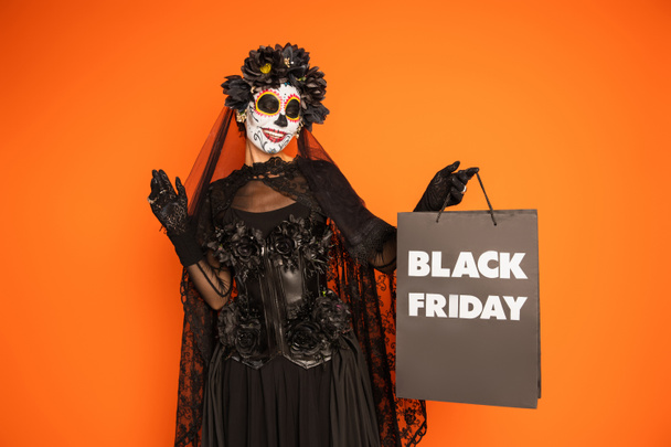 happy woman in traditional santa muerte makeup and costume holding black friday shopping bag isolated on orange - Foto, Bild