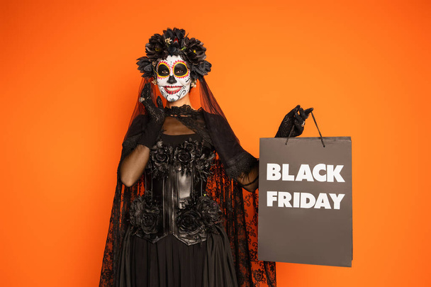 happy woman in mexican day of dead makeup and costume holding black friday shopping bag isolated on orange - Фото, изображение