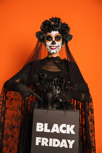 cheerful woman in spooky halloween makeup and witch costume holding black friday shopping bag isolated on orange - Foto, Bild