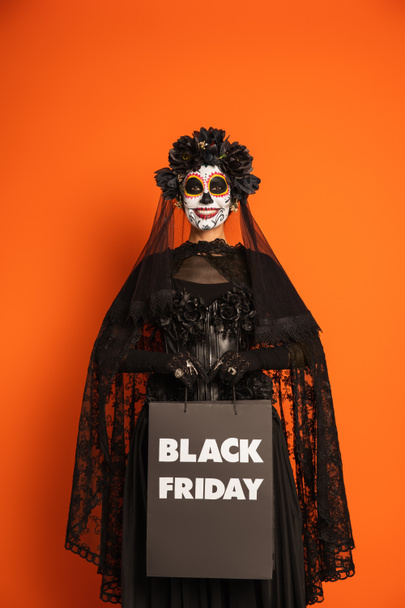 sorrindo mulher em assustador halloween maquiagem e traje segurando preto sexta-feira saco de compras isolado em laranja - Foto, Imagem