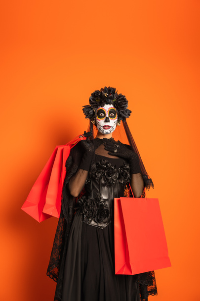 astonished woman in catrina makeup and black creepy costume holding shopping bags and touching chest on orange background - Foto, Bild