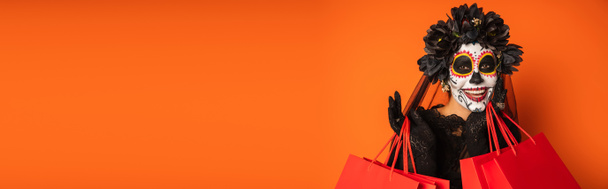 glückliche Frau in schwarzem Kranz und gruseligem Halloween-Make-up, die in die Kamera neben Einkaufstüten schaut, vereinzelt auf orangefarbenem Banner - Foto, Bild