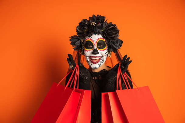 glimlachende vrouw in zwart Halloween kostuum en suiker schedel make-up houden boodschappentassen en kijken naar de camera op oranje achtergrond - Foto, afbeelding