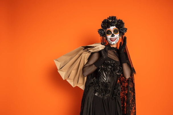 happy woman in mexican day of dead makeup and black costume standing with shopping bags on orange background - Photo, Image