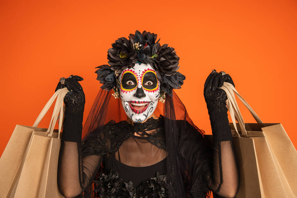 amazed woman in black halloween costume and creepy makeup holding shopping bags isolated on orange - Foto, imagen
