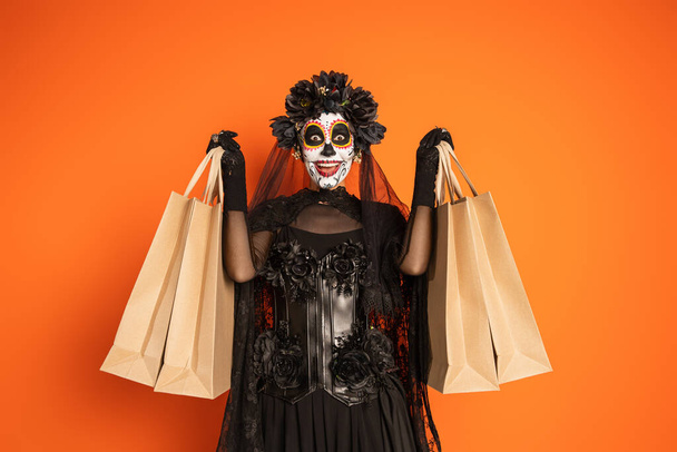 ενθουσιασμένοι γυναίκα σε μαύρο αποκριάτικο κοστούμι με στεφάνι και τρομακτικό μακιγιάζ κρατώντας τσάντες ψώνια απομονώνονται σε πορτοκαλί - Φωτογραφία, εικόνα