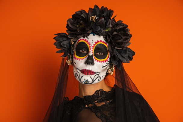 retrato de la mujer en corona negra y el cráneo de azúcar de Halloween maquillaje mirando a la cámara aislada en naranja - Foto, imagen