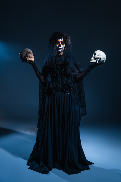 Siyah kostümlü ve korkutucu cadılar bayramı makyajlı bir kadın. Kafataslarını koyu renk arka planda mavi ışıklı tutuyor. - Fotoğraf, Görsel