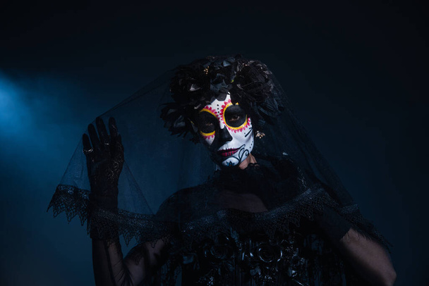 mujer en tradicional santa muerte maquillaje tocando velo negro y mirando a la cámara sobre fondo oscuro con luz azul - Foto, Imagen