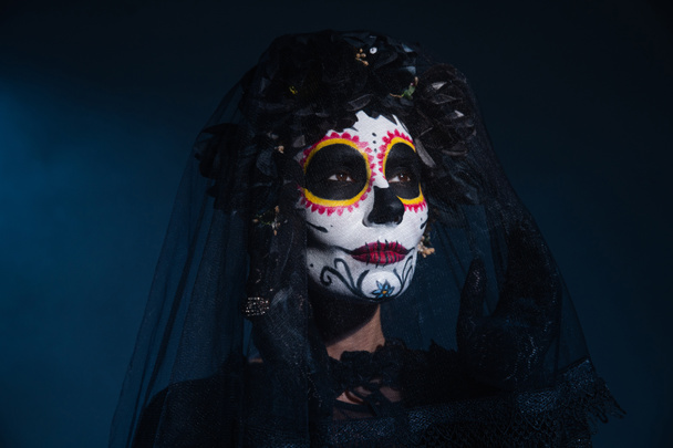 donna in tradizionale messicano trucco catrina toccare velo e guardando lontano su sfondo blu scuro - Foto, immagini