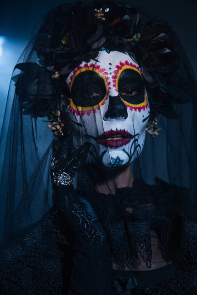 retrato de la mujer en el maquillaje de Halloween espeluznante mirando a la cámara bajo el velo negro sobre fondo oscuro con luz azul - Foto, Imagen