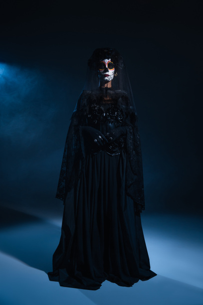 longitud completa de la mujer en el maquillaje tradicional de santa muerte y vestido negro de pie en luz azul sobre fondo oscuro - Foto, Imagen