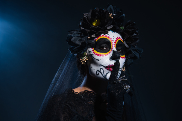 woman in spooky halloween makeup and black wreath with veil showing hush sign on dark blue background - Photo, Image