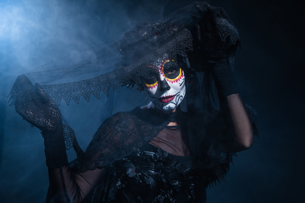mujer en el maquillaje de Halloween miedo sosteniendo velo de encaje negro y mirando a la cámara en el fondo oscuro con humo azul - Foto, Imagen
