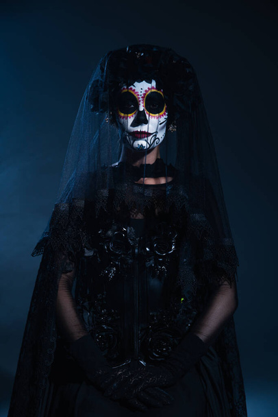 mujer en tradicional mexicana santa muerte maquillaje y traje mirando a la cámara sobre fondo azul oscuro - Foto, imagen