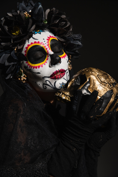 woman with scary halloween makeup and closed eyes holding golden skull isolated on black - Fotó, kép
