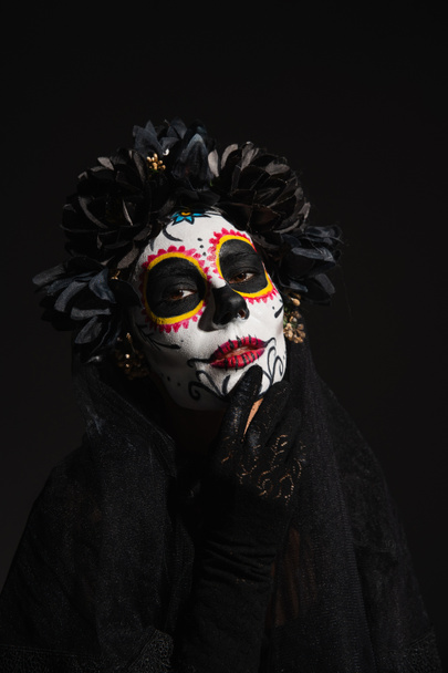 γυναίκα σε σκούρο στεφάνι αγγίζοντας το πρόσωπο με παραδοσιακό μακιγιάζ santa muerte κοιτάζοντας κάμερα απομονωμένη σε μαύρο - Φωτογραφία, εικόνα