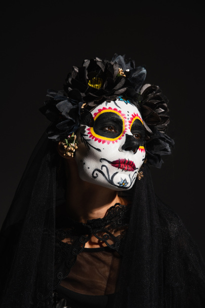 portrait of woman with creepy halloween makeup and dark wreath looking away isolated on black - Photo, Image