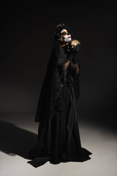 full length of woman in black witch costume and traditional santa muerte makeup standing with golden skull on dark background - Fotó, kép