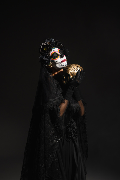 woman with sugar skull makeup wearing dark costume with veil and holding golden skull isolated on black - Photo, Image