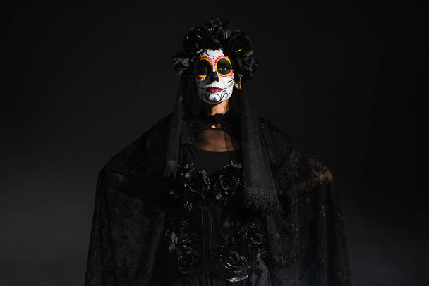 γυναίκα σε τρομακτικό αποκριάτικο κοστούμι και μακιγιάζ στέκεται με τα χέρια πίσω από την πλάτη απομονώνονται σε μαύρο - Φωτογραφία, εικόνα