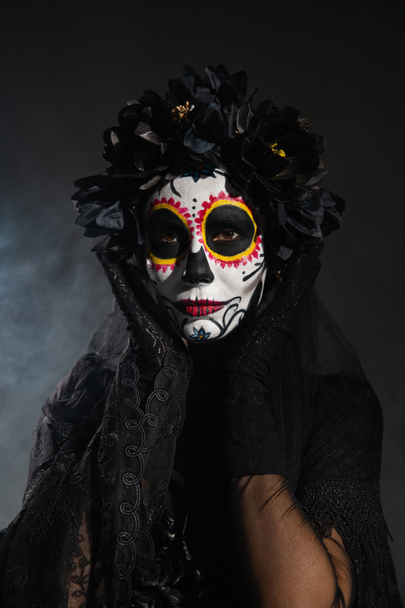 woman in black wreath holding hands near face with sugar skull makeup on dark smoky background - Photo, Image