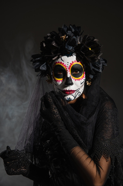 woman in black wreath and traditional santa muerte makeup looking at camera on dark foggy background - Fotografie, Obrázek