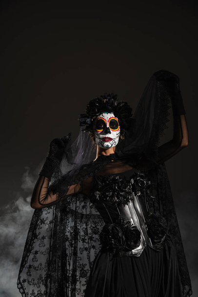 woman in traditional mexican day of dead makeup posing with black lace veil on dark smoky background - Foto, Imagem