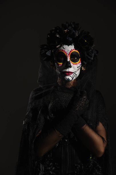 woman with mexican day of dead makeup wrapping herself in dark veil and looking at camera isolated on black - Foto, Imagem