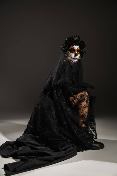 volledige lengte van de vrouw met suiker schedel make-up dragen van zwarte jurk met kant sluier poseren op donkere achtergrond - Foto, afbeelding