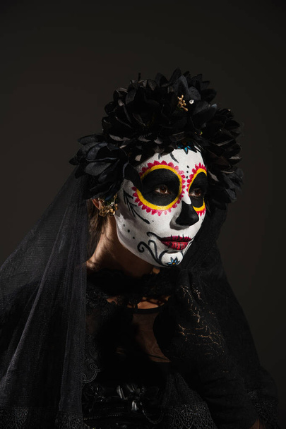 woman in dark halloween costume and sugar skull makeup looking away isolated on black - Fotó, kép