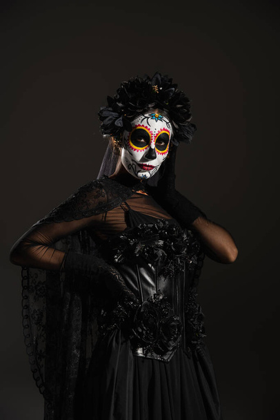 vrouw in donker halloween kostuum en suiker schedel make-up poseren met de hand op taille geïsoleerd op zwart - Foto, afbeelding