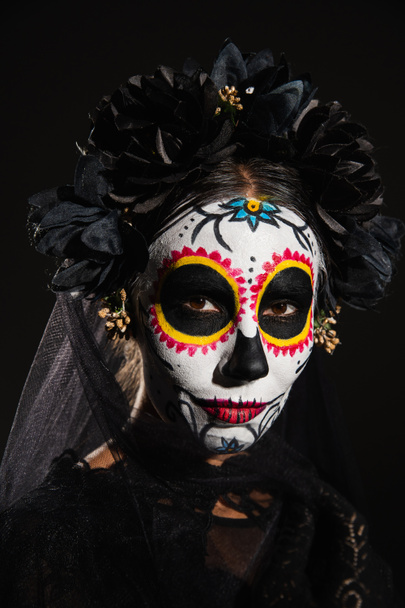 portrait of woman in halloween sugar skull makeup and dark wreath with veil looking at camera isolated on black - Фото, зображення