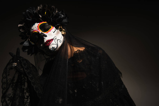 mujer en espeluznante maquillaje de halloween y corona oscura con velo de encaje posando con los ojos cerrados sobre fondo oscuro - Foto, imagen