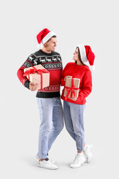 Beautiful happy couple in Santa hats holding Christmas gifts on light background - Fotoğraf, Görsel