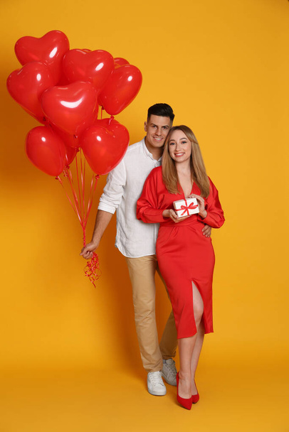 Lovely couple with heart shaped balloons and gift on yellow background. Valentine's day celebration - Φωτογραφία, εικόνα