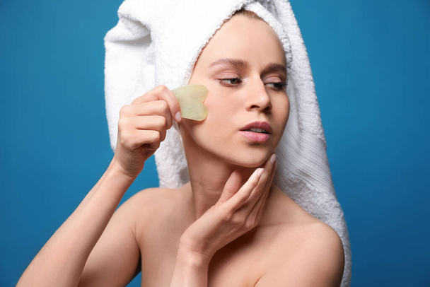 Beautiful young woman doing facial massage with gua sha tool on blue background - Zdjęcie, obraz