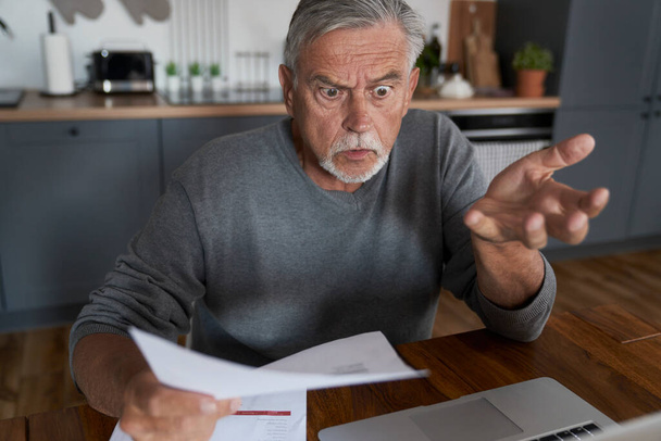 Shocked senior man looking at laptop and holding documents in hand - Photo, Image