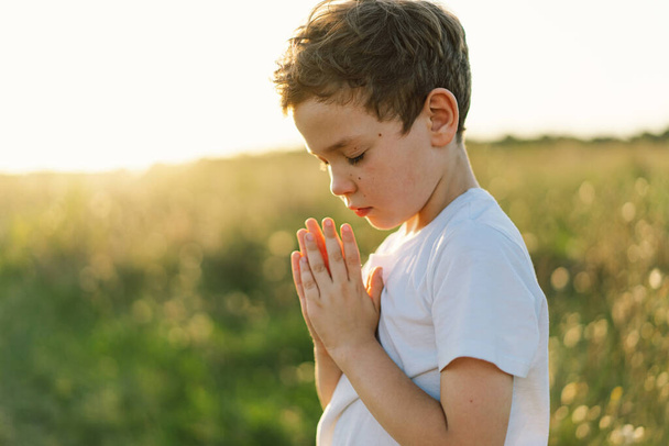 Мальчик закрыл ей глаза и молился в поле на закате. Руки сложены в молитвенной концепции веры, духовности и религии. - Фото, изображение